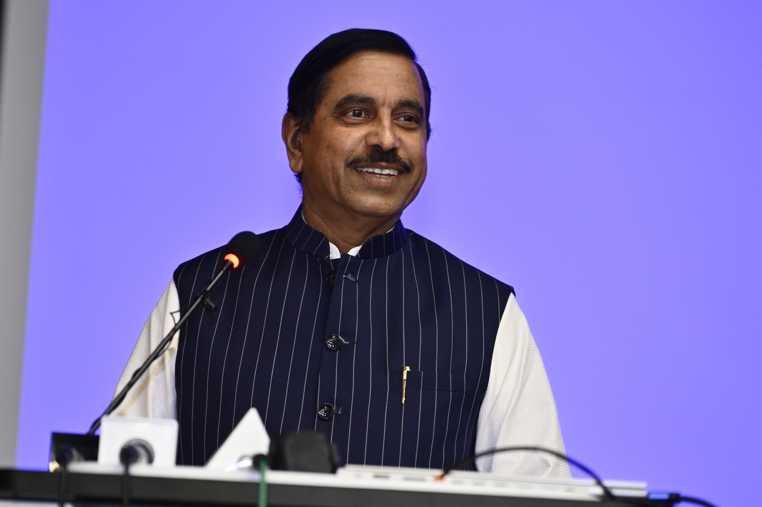 Professional keynote speaker smiling while delivering a speech at a corporate conference with a blue-lit background.