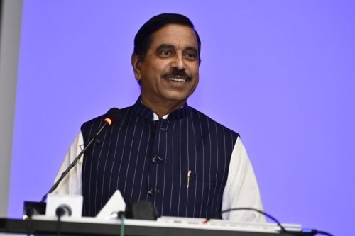 Professional keynote speaker smiling while delivering a speech at a corporate conference with a blue-lit background.