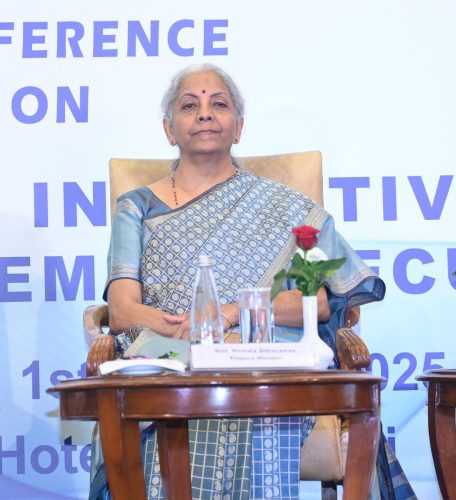 India's Finance Minister seated on stage during an official conference with a conference backdrop, water bottle, glass, and red rose on the table in front.