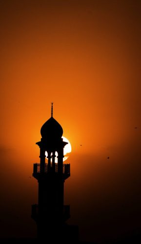 Silhouette of a mosque minaret with the setting sun behind it against a vibrant orange and red sky, with birds flying across the horizon.