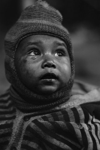 Black-and-white close-up portrait of a child with a tear on the cheek, looking upward with expressive eyes in soft natural light.